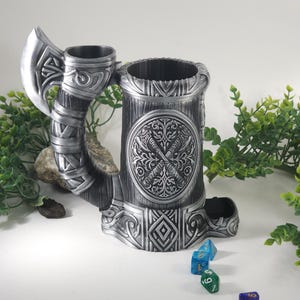 May include: A silver metal dice tower shaped like a Viking drinking horn with an axe handle. The tower has an intricate design with a circular emblem featuring crossed axes. The tower is sitting on a white surface with four dice scattered around it.