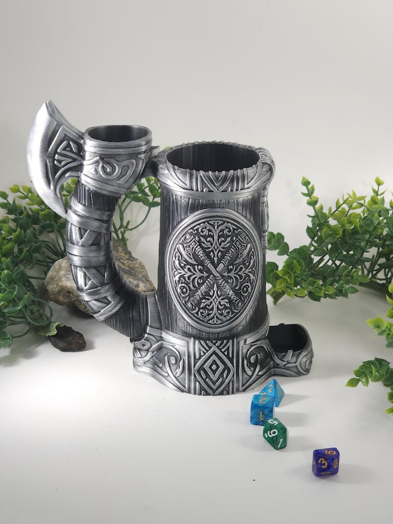 May include: A silver metal dice tower shaped like a Viking drinking horn with an axe handle. The tower has an intricate design with a circular emblem featuring crossed axes. The tower is sitting on a white surface with four dice scattered around it.
