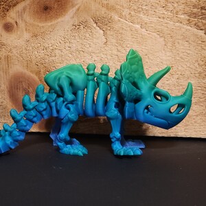 May include: A 3D printed skeleton of a triceratops dinosaur, with a gradient of blue and green colors. The dinosaur is standing on a wooden surface.