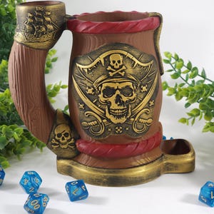 May include: A brown ceramic pirate-themed dice tower with a skull and crossbones design. The tower has a red rope detail and a gold trim. There are blue dice on the table in front of the tower.
