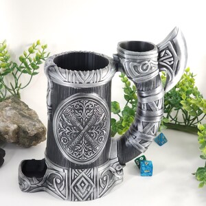 May include: A silver metal dice tower shaped like a mug with an axe handle. The tower has a circular design with two crossed swords and a floral pattern. The mug has a compartment for dice and a separate compartment for holding dice.