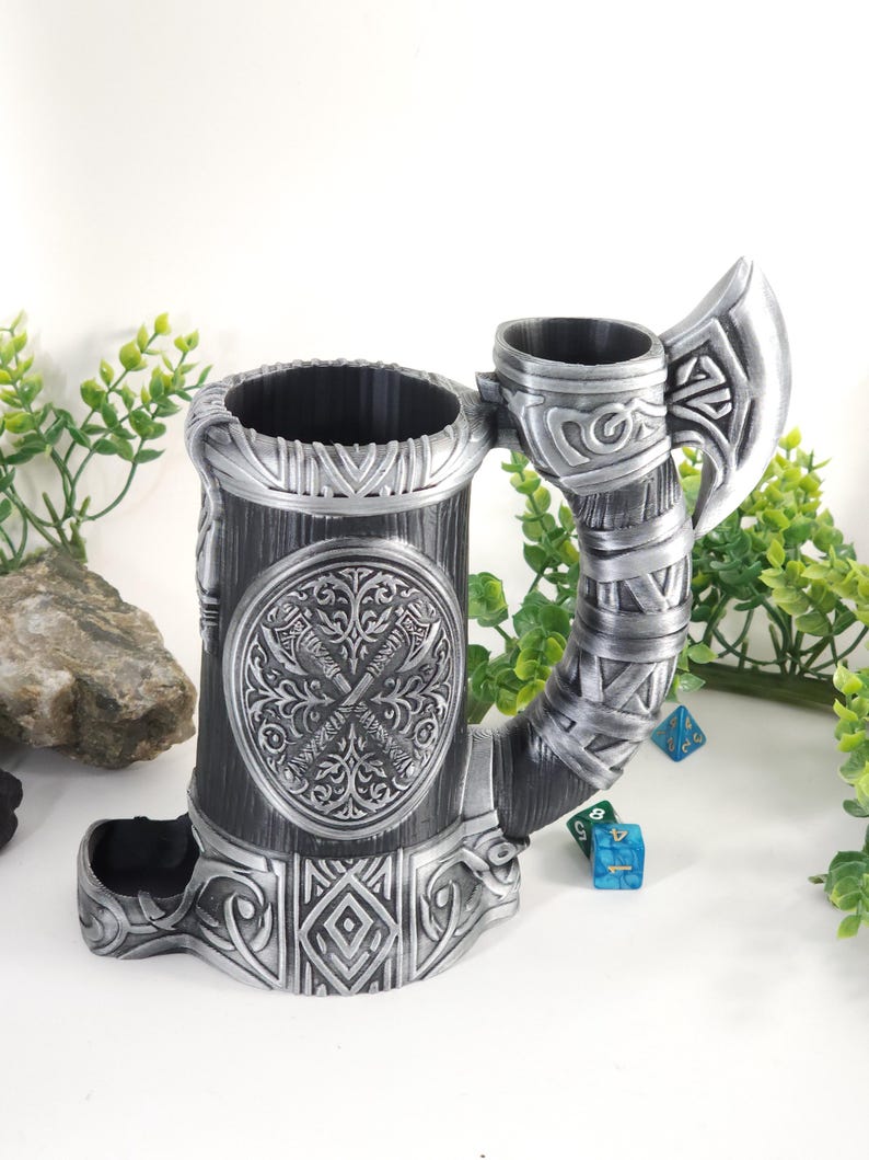 May include: A silver metal dice tower shaped like a mug with an axe handle. The tower has a circular design with two crossed swords and a floral pattern. The mug has a compartment for dice and a separate compartment for holding dice.