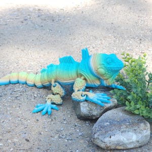 May include: A blue and green iguana figurine with articulated limbs, posed on two rocks. The figurine is made of plastic and has a realistic appearance.