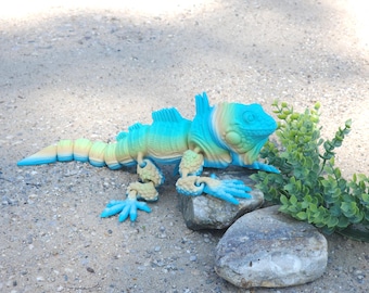 May include: A blue and green iguana figurine with articulated limbs, posed on two rocks. The figurine is made of plastic and has a realistic appearance.