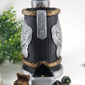 May include: A black and silver dice tower with a skull and crossbones design. The tower has a gold trim and a compartment for dice. There are several green dice with gold numbers on a white surface.
