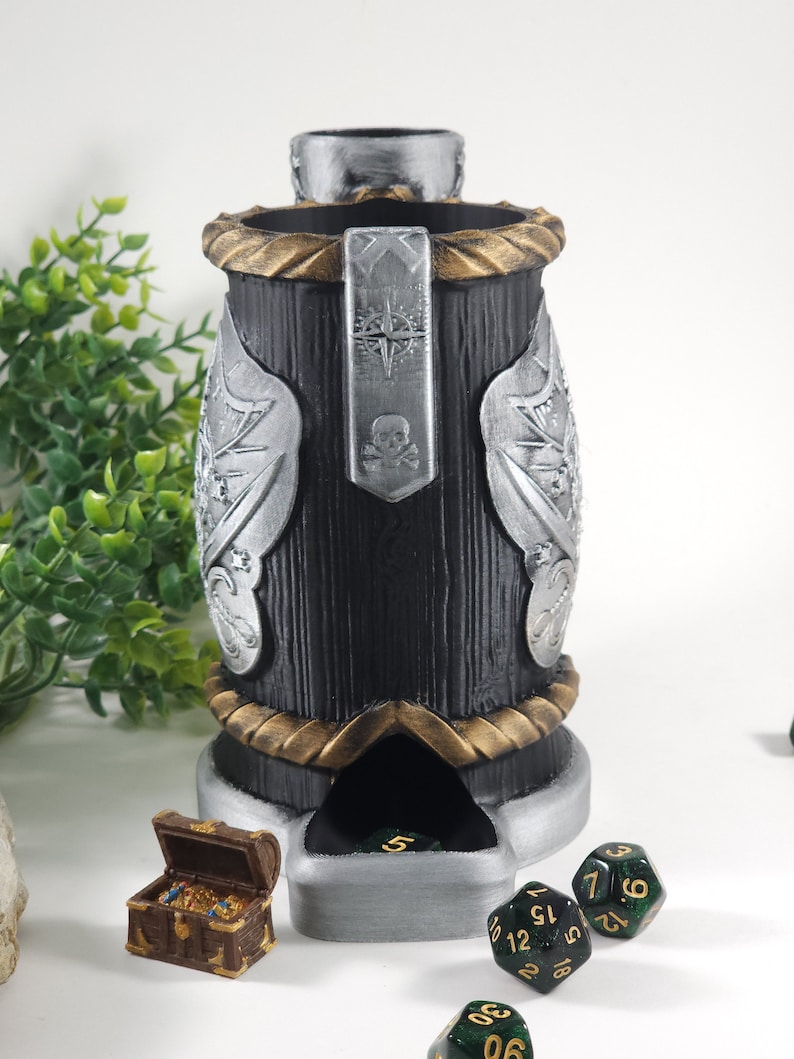 May include: A black and silver dice tower with a skull and crossbones design. The tower has a gold trim and a compartment for dice. There are several green dice with gold numbers on a white surface.