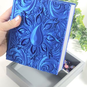 May include: A blue, ornate box with a raised design of a teardrop and floral pattern. The box is open, revealing a gray interior with a compartment for dice.