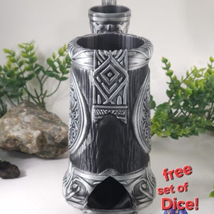 May include: A silver metal dice tower with intricate carvings and a slot for rolling dice. The tower has a small lid on top. A set of colorful dice are scattered on a white surface in front of the tower. The text "free set of Dice!" is printed on a white background.