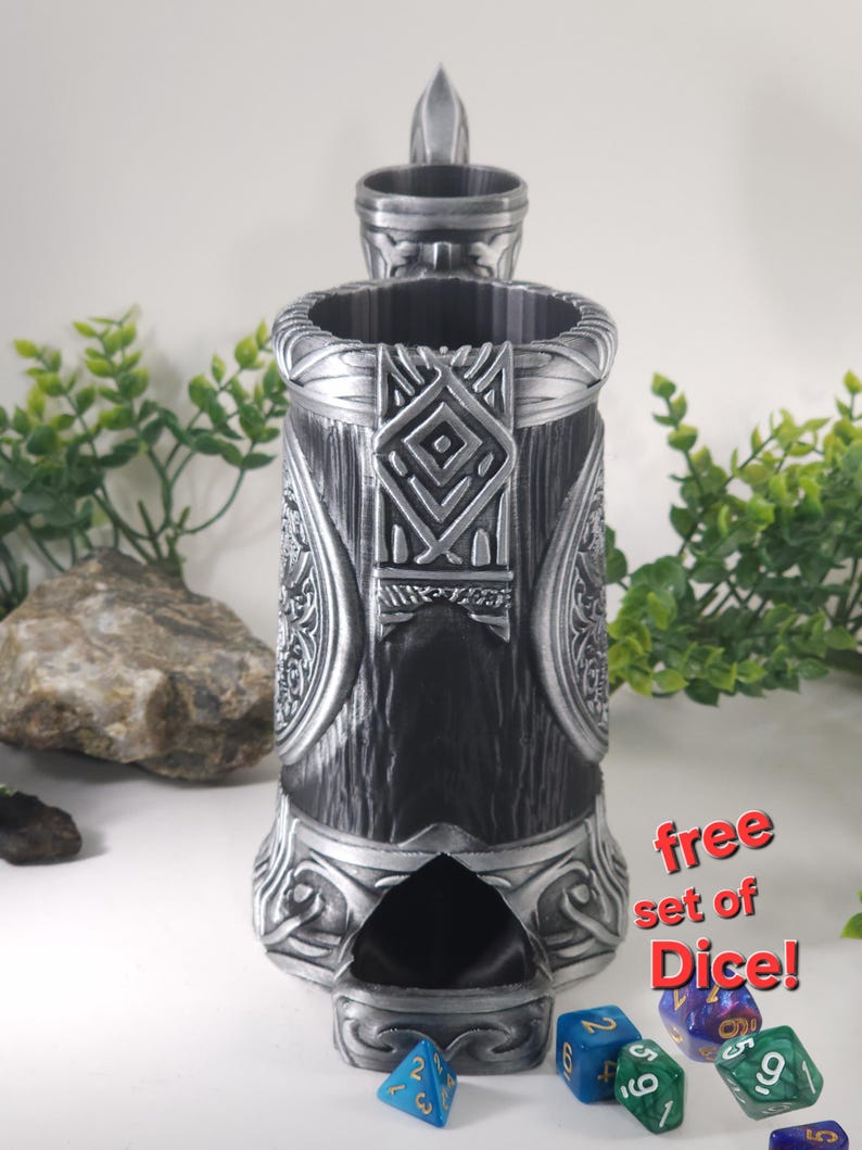 May include: A silver metal dice tower with intricate carvings and a slot for rolling dice. The tower has a small lid on top. A set of colorful dice are scattered on a white surface in front of the tower. The text "free set of Dice!" is printed on a white background.