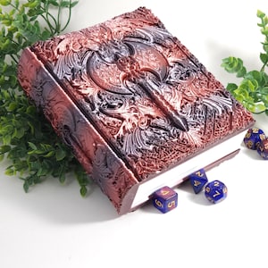May include: A dark brown leather-bound journal with an embossed axe design. The journal is closed and lying on a white surface with several blue and gold dice scattered around it.