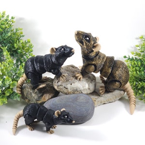 May include: Three articulated toy rats, one black, one brown, and one black with a tan tail, are posed on rocks. The rats have realistic details and are made of plastic.