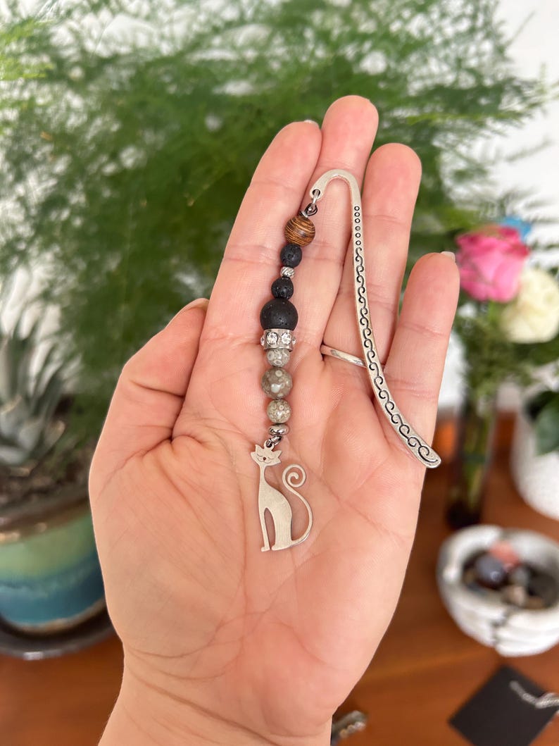 May include: A silver cat bookmark with a beaded strand of brown, black, and grey beads. The bookmark has a curved top with decorative cutouts. The cat charm is a simple silhouette.