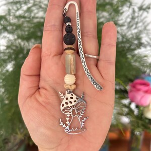 May include: A handmade bookmark with a silver mushroom charm and decorative beads. The beads include black lava stones, light brown and beige wooden beads. The bookmark is held in a hand against a background of green foliage and a pink rose.