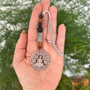 May include: A silver bookmark with a tree of life pendant and a beaded strand. The pendant features a seated figure within a tree design. The beaded strand includes black, brown, and blue-green beads. The bookmark is held in a hand.
