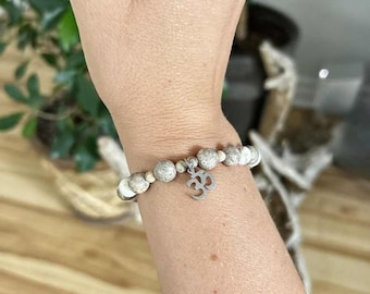 May include: A beaded bracelet with a silver Om charm. The bracelet features light beige and white beads. The charm is a stylized Om symbol. The bracelet is worn on a wrist.