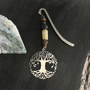 May include: Silver metal bookmark with a tree of life pendant. The bookmark features a decorative design with a curved top and a series of beads in shades of brown, black, and cream. The pendant is a detailed cutout of a tree.