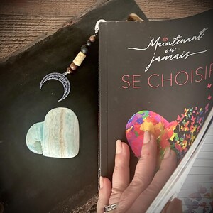 May include: A bookmark featuring a crescent moon charm, wooden beads, and a heart-shaped stone. The bookmark is in a book with the French title "Maintenant ou jamais SE CHOISIR". The heart-shaped stone is a pale blue and white colour.