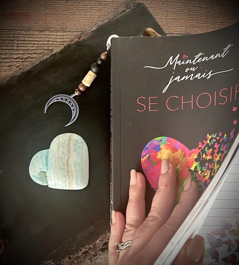 May include: A bookmark featuring a crescent moon charm, wooden beads, and a heart-shaped stone. The bookmark is in a book with the French title "Maintenant ou jamais SE CHOISIR". The heart-shaped stone is a pale blue and white colour.