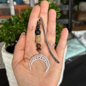 May include: A silver bookmark featuring a crescent moon design, adorned with beads in shades of black, brown, and tan. The bookmark is held in a hand, with a green plant visible in the background. The beads have different textures.