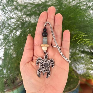 May include: A silver metal bookmark featuring a turtle pendant. The pendant is connected to a beaded strand with black, blue, and cream beads. The bookmark is held in a hand against a backdrop of green leaves.