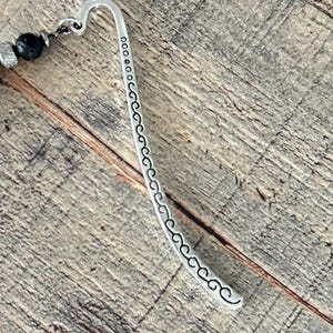 May include: A silver metal bookmark with a curved hook design and a decorative pattern along the curved edge. The bookmark is connected to a strand of black beads and silver accents. The background is a wooden surface.