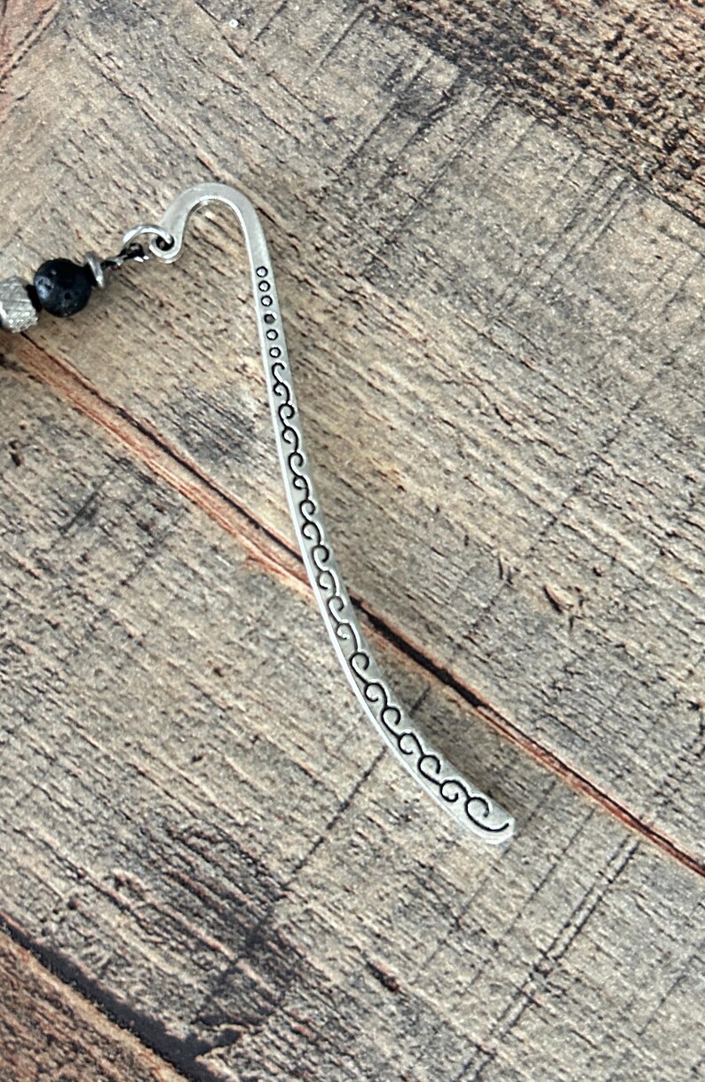 May include: A silver metal bookmark with a curved hook design and a decorative pattern along the curved edge. The bookmark is connected to a strand of black beads and silver accents. The background is a wooden surface.