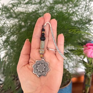 May include: A silver bookmark with a floral pendant. The pendant is an eight-pointed star design. The bookmark has a beaded accent with black and gold beads. The background features a green plant. The bookmark is a decorative item.