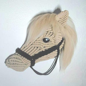 Może przedstawiać: Beżowa makrama głowa konia do zawieszenia na ścianie z czarnym uzdą i płynącą blond grzywą. Głowa konia wykonana jest ze sznurka, z wyraźnym okiem i uchem. Unikalny, ręcznie robiony element.