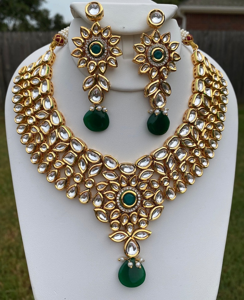 Peut inclure: Un collier et des boucles d'oreilles en or avec un pendentif en pierre verte et de nombreuses pierres claires. Le collier est un design superpos&eacute; avec un grand groupe de pierres en forme de fleur au centre.