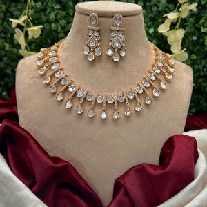 May include: A gold-toned necklace and matching earrings set. The jewelry features clear, teardrop-shaped stones and smaller, round stones. The necklace and earrings are displayed on a mannequin with a burgundy and white fabric backdrop.