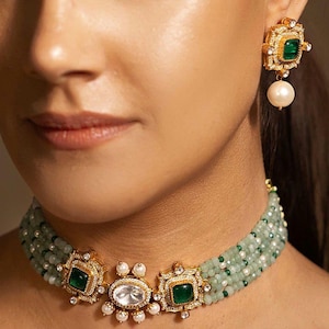 Może przedstawiać: Zbliżenie złotego zestawu biżuterii z naszyjnikiem typu choker z koralików i pasującymi kolczykami. Naszyjnik ma zielone koraliki, złote akcenty i kwadratowe kamienie w kolorze szmaragdu. Kolczyki mają podobny design z perłową zawieszką.