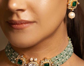 Peut inclure: Gros plan sur un ensemble de bijoux dorés comprenant un collier ras du cou à perles et des boucles d'oreilles assorties. Le collier est orné de perles vertes, d'accents dorés et de pierres carrées de couleur émeraude. Les boucles d'oreilles ont un design similaire avec une perle.