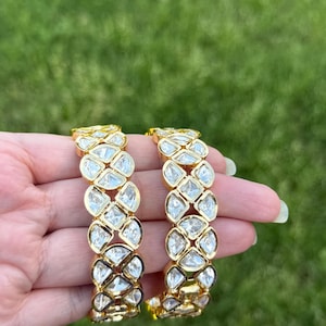 May include: Two gold-colored bangles with clear, faceted stones. The stones are set in a repeating pattern along the length of the bangles. The background is a blurred green lawn.
