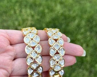 Peut inclure: Deux bracelets dorés ornés de pierres claires et facettées. Les pierres sont disposées en motif répétitif sur toute la longueur des bracelets. L'arrière-plan est une pelouse verte floue.