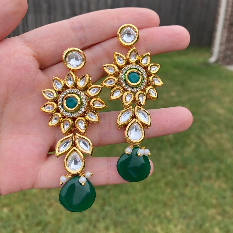 Peut inclure: Boucles d'oreilles pendantes dor&eacute;es avec une pierre verte et des strass transparents. Les boucles d'oreilles ont un motif floral et sont rehauss&eacute;es de perles blanches.