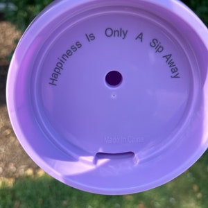 Puede incluir: Tapa de pl&aacute;stico lila con el texto "Happiness Is Only A Sip Away" dispuesto en c&iacute;rculo. La tapa tiene un peque&ntilde;o agujero central y una ranura. Tambi&eacute;n se puede leer "Made In China".
