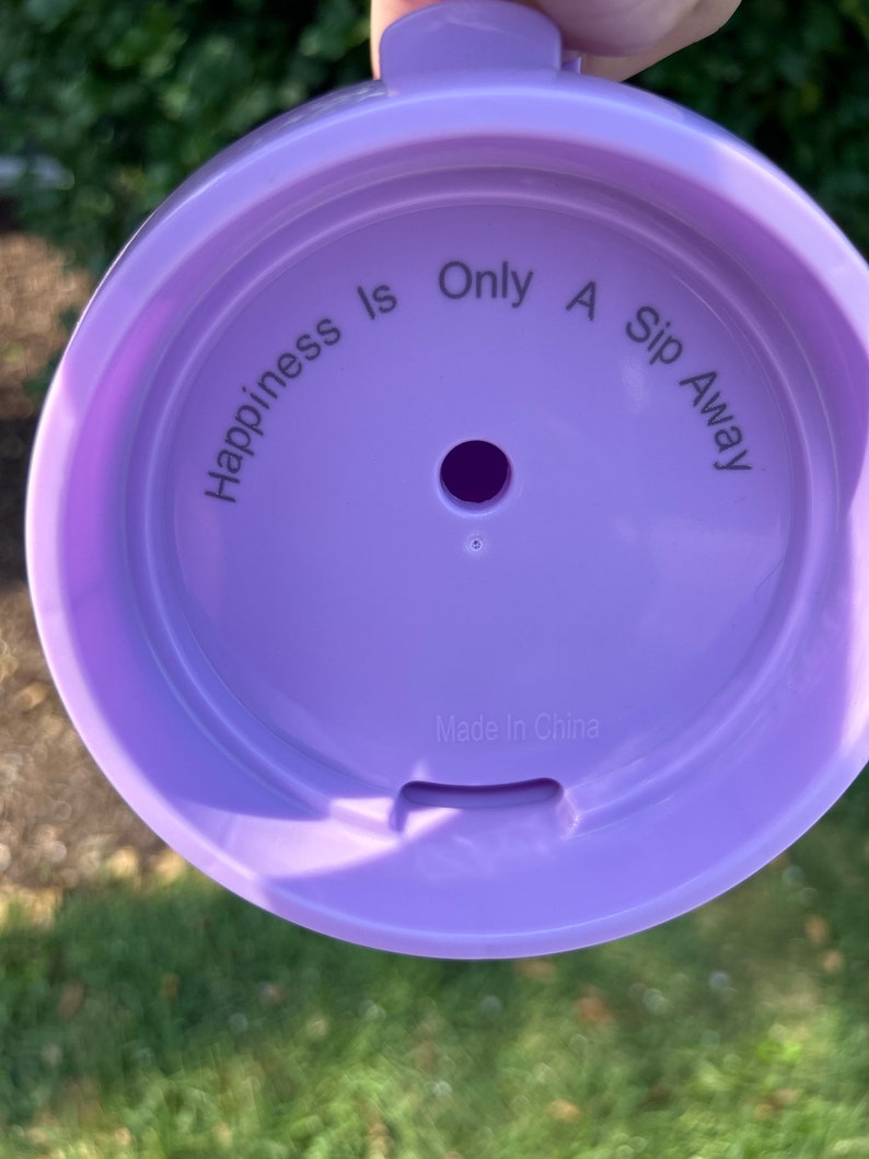 Puede incluir: Tapa de pl&aacute;stico lila con el texto "Happiness Is Only A Sip Away" dispuesto en c&iacute;rculo. La tapa tiene un peque&ntilde;o agujero central y una ranura. Tambi&eacute;n se puede leer "Made In China".