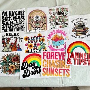 Puede incluir: Una colecci&oacute;n de pegatinas cuadradas con varios dise&ntilde;os y texto. Los dise&ntilde;os incluyen un chef esqueleto, una mariposa con un arco&iacute;ris, un fuego de basurero, una pila de libros, un arco&iacute;ris y frases como "Forever Chasing Sunsets" y "Tanned & Tipsy."