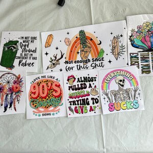 Puede incluir: Una colecci&oacute;n de pegatinas coloridas con varios dise&ntilde;os y texto. Una pegatina muestra un mapache en un cubo de basura con texto. Otras pegatinas incluyen un arco&iacute;ris, un esqueleto y un atrapasue&ntilde;os. Las pegatinas tienen una variedad de fuentes y colores.