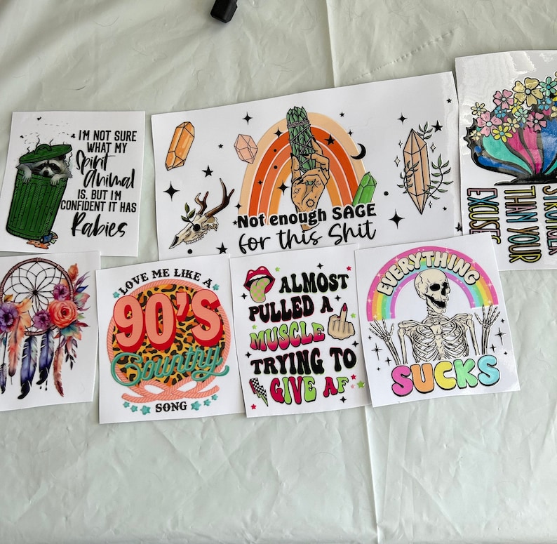 Puede incluir: Una colecci&oacute;n de pegatinas coloridas con varios dise&ntilde;os y texto. Una pegatina muestra un mapache en un cubo de basura con texto. Otras pegatinas incluyen un arco&iacute;ris, un esqueleto y un atrapasue&ntilde;os. Las pegatinas tienen una variedad de fuentes y colores.