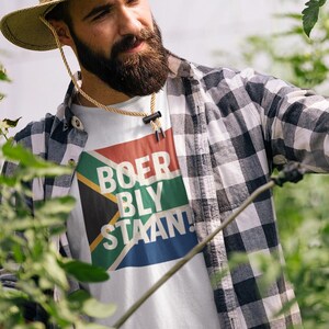 Peut inclure: Un homme portant un chapeau de paille et une chemise à carreaux s'occupe de plants de tomates dans une serre. Le t-shirt blanc porte l'inscription "BOER BLY STAAN!" devant les couleurs du drapeau sud-africain. Les plantes ont des tomates vertes.
