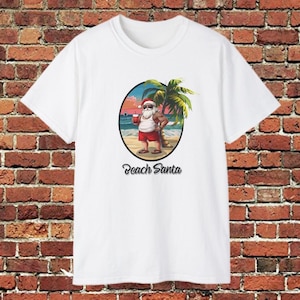 May include: White t-shirt featuring a cartoon illustration of Santa Claus on a beach, holding a drink. Santa is wearing sunglasses, red shorts, and sandals. The text "Beach Santa" is below the image.