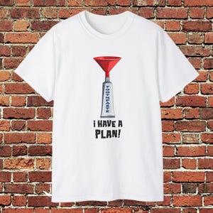 Puede incluir: Camiseta blanca con un embudo rojo sobre una ilustración de un tubo de pasta de dientes. La palabra "TOOTHPASTE" está impresa verticalmente en el tubo, y debajo, la frase "I HAVE A PLAN!" se muestra en negro.