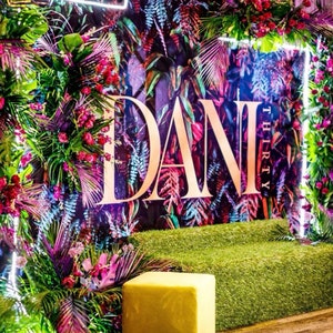 K&ouml;nnte beinhalten: Ein tropischer Hintergrund mit &uuml;ppigem Gr&uuml;n und leuchtenden Blumen. Der Hintergrund zeigt den Namen "Dani Thirty" in goldenen Buchstaben. Vor dem Hintergrund befindet sich eine grasartige Oberfl&auml;che in Gr&uuml;n, mit einem quadratischen Ottoman in Gelb.