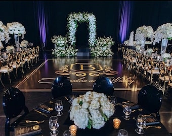 May include: A formal event space featuring a black dance floor with a gold monogrammed letter B and the text Angelica and Wesley. The room includes round tables with black chairs, white floral centerpieces, and a large white floral arch.