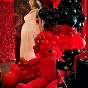 K&ouml;nnte beinhalten: Ein roter Teddyb&auml;r sitzt vor einem Hintergrund aus roten und schwarzen Luftballons. Eine schwangere Frau in einem rosa Kleid steht hinter den Ballons.