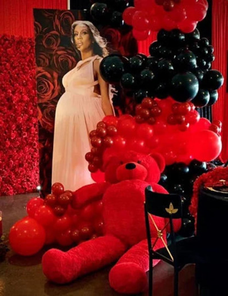 K&ouml;nnte beinhalten: Ein roter Teddyb&auml;r sitzt vor einem Hintergrund aus roten und schwarzen Luftballons. Eine schwangere Frau in einem rosa Kleid steht hinter den Ballons.