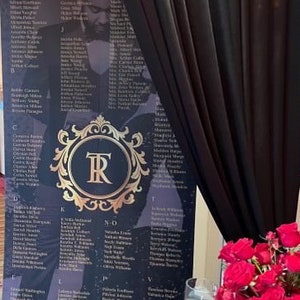 May include: A black and gold wedding seating chart with a gold monogram of the letters "TR" in a decorative frame. The chart lists guest names in alphabetical order.