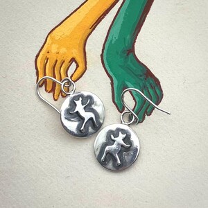 Könnte beinhalten: Ein Paar silberne Ohrringe mit einem kreisförmigen Design, das eine stilisierte Tierfigur zeigt. Die Ohrringe haben einen Hakenverschluss. Der Hintergrund zeigt gemalte Hände in Gelb und Grün.