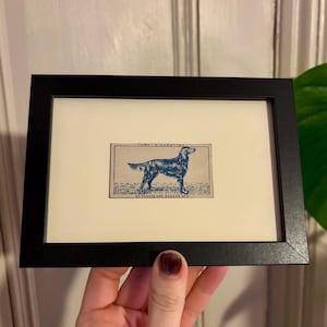 May include: A small rectangular black frame containing a vintage-style print of an Irish Setter dog in blue ink on a cream background. The print features the text "TURF CIGARETTES" at the top and "IRISH SETTER 50 FAMOUS DOG BREEDS N°6" at the bottom.